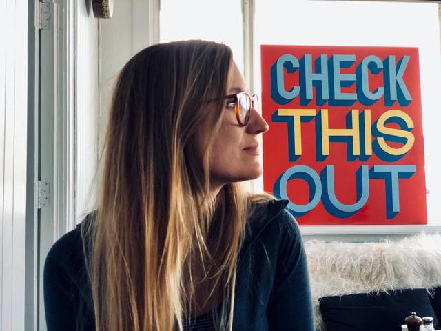 lady with long hair and glasses in a shop with a sign in the window Check This Out