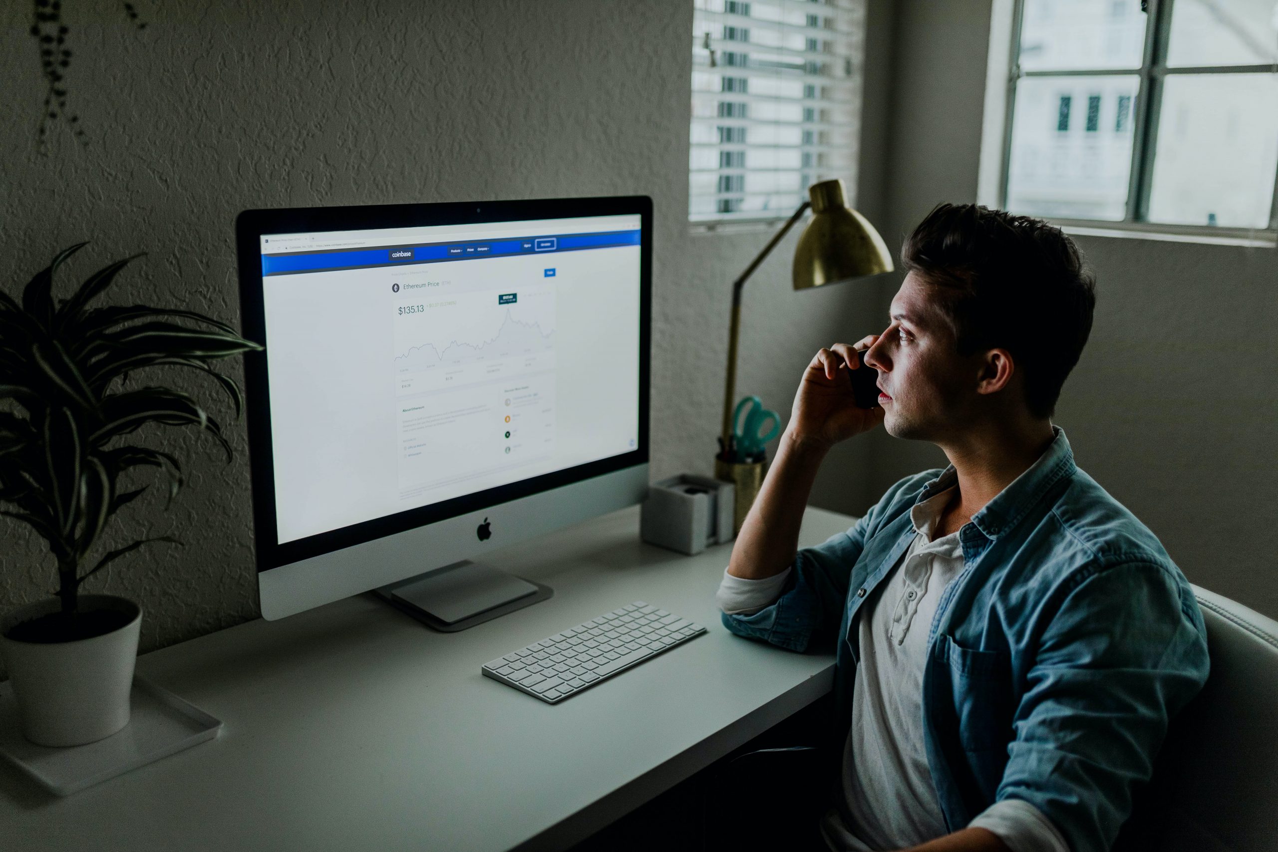 A desk with a young man sat on a chair with his hand up to his face looking at a computer