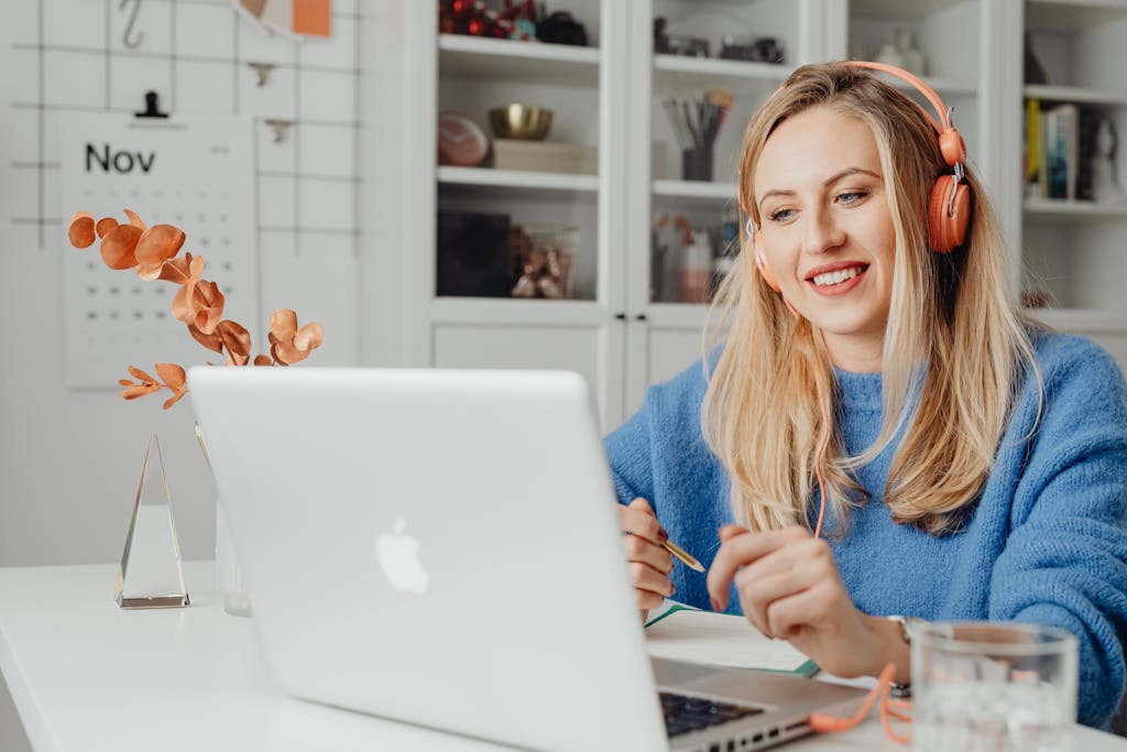Topics 4 Blonde woman smiling while working remotely on a laptop, wearing headphones in a cozy home office.