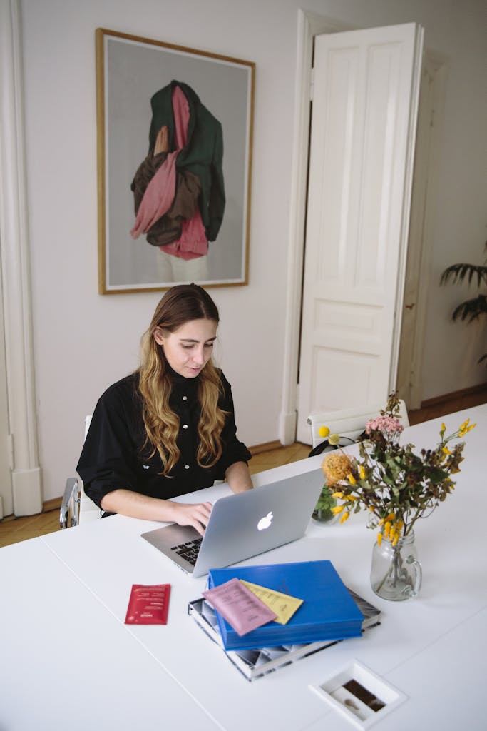 Topics 1 Young woman using a laptop at a desk with documents and flowers, in a bright, modern workspace.